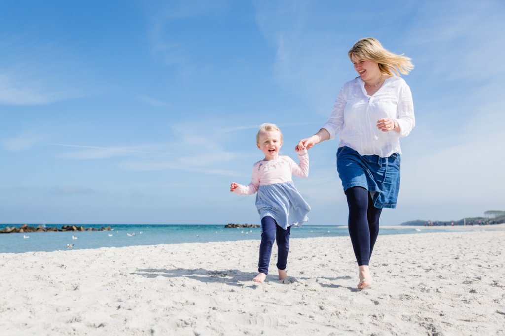Tochter flitzt mit Mama an der Hand am Strand entlang.